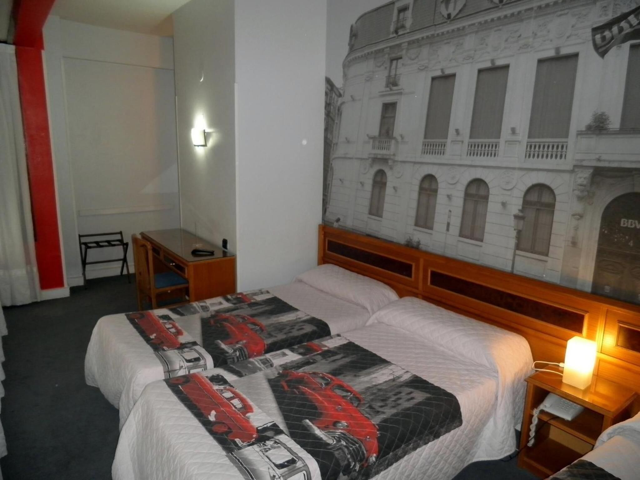 A hotel room with a bed, a desk, a chair, a nightstand with a lamp, and a wall with a building mural.