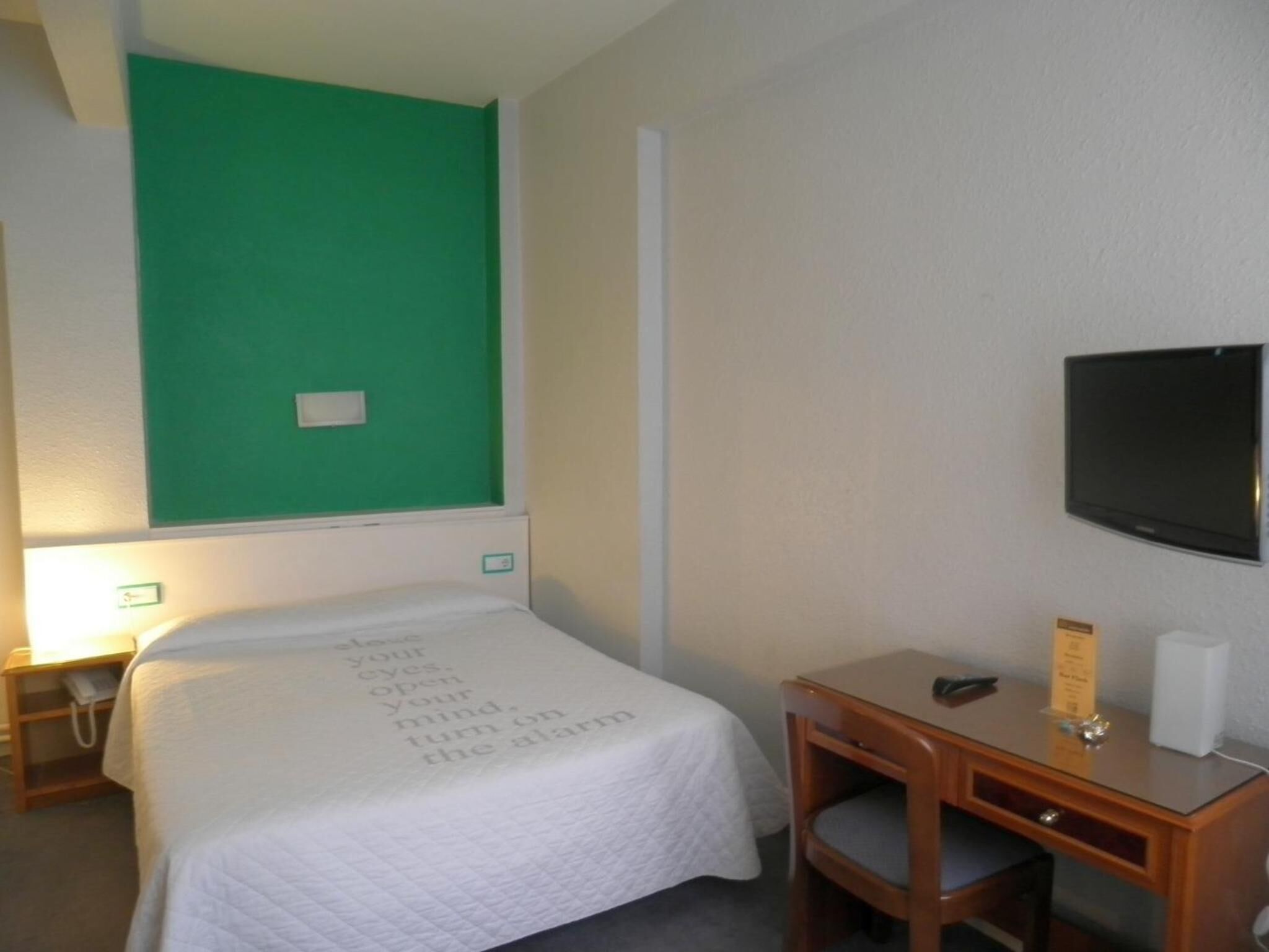 Habitación de hotel con cama, escritorio, silla, televisión y pared verde.