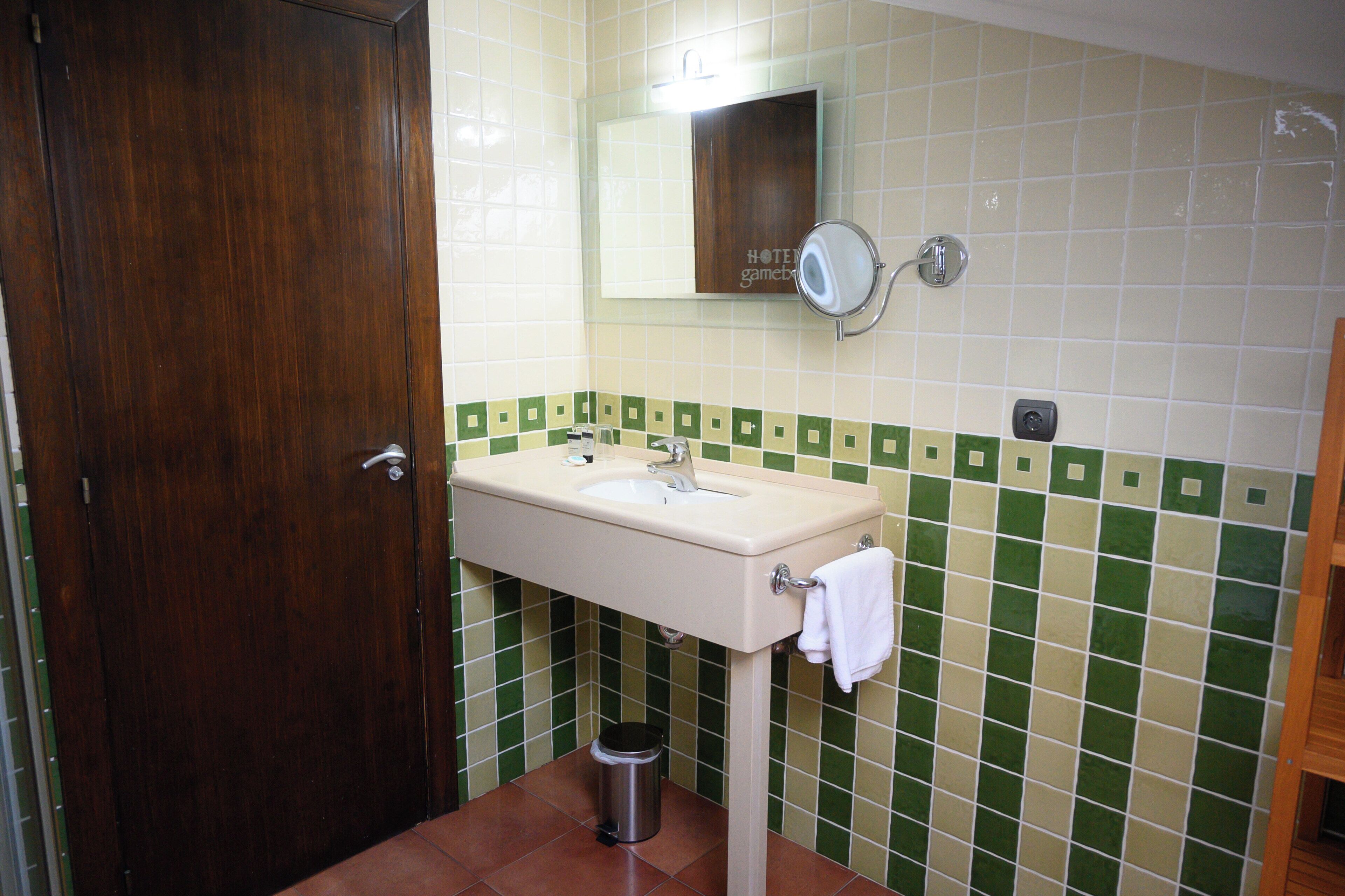 Een badkamer met een houten deur, een wastafel met spiegel en groene en witte betegelde muren.