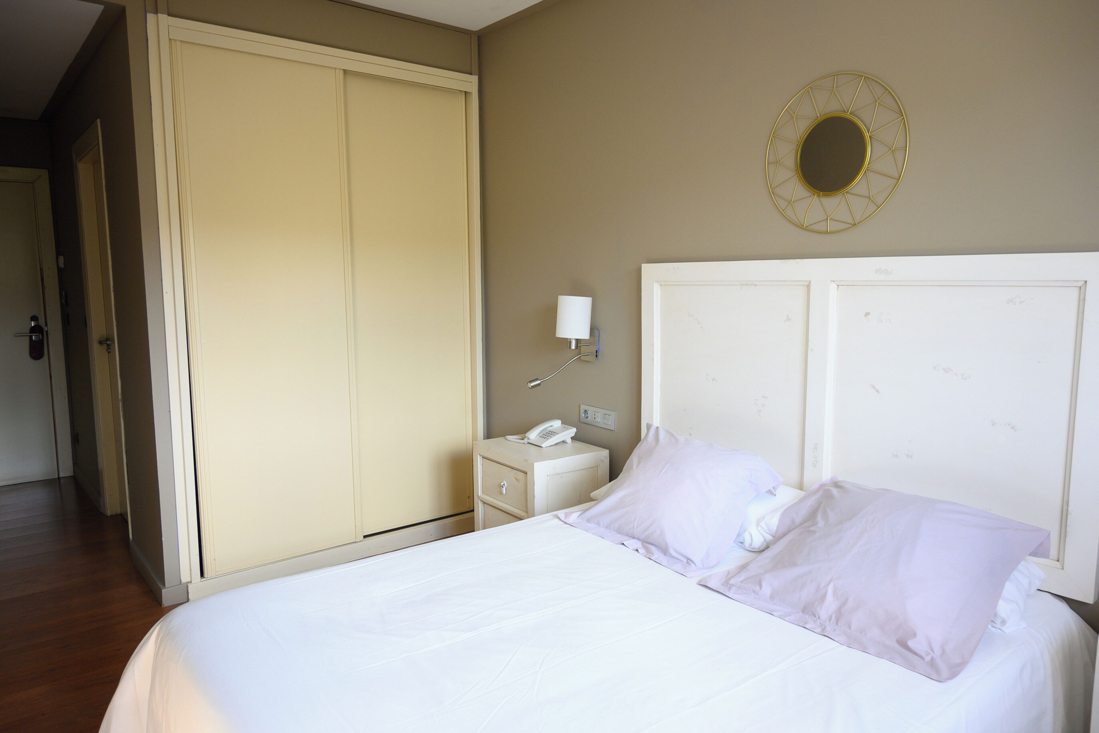 A hotel room with a bed, a bedside table with a phone, a wall-mounted light, and a wardrobe.