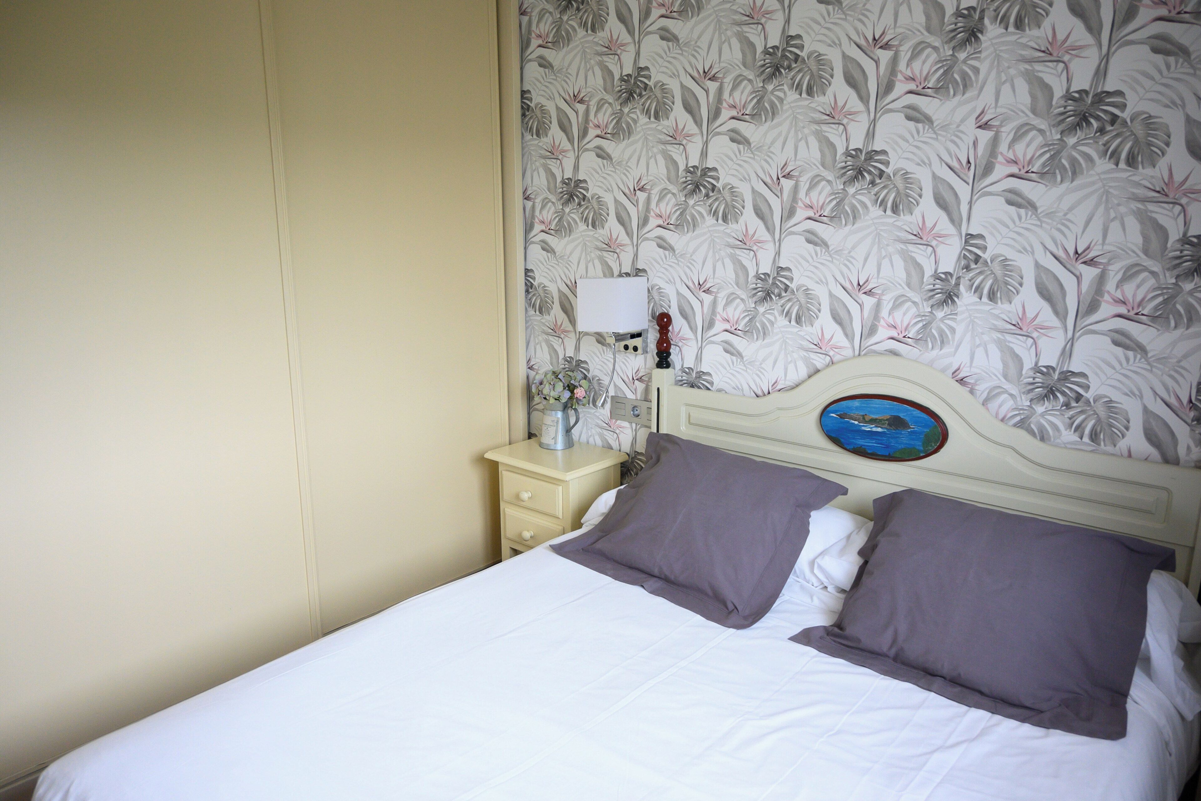 A bed with white bedding and two pillows, a nightstand with a lamp and a small vase of flowers, and a wall with a floral wallpaper pattern.