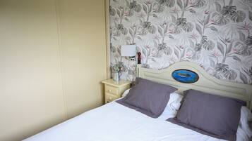 A bed with white bedding and two pillows, a nightstand with a lamp and a small vase of flowers, and a wall with a floral wallpaper pattern.