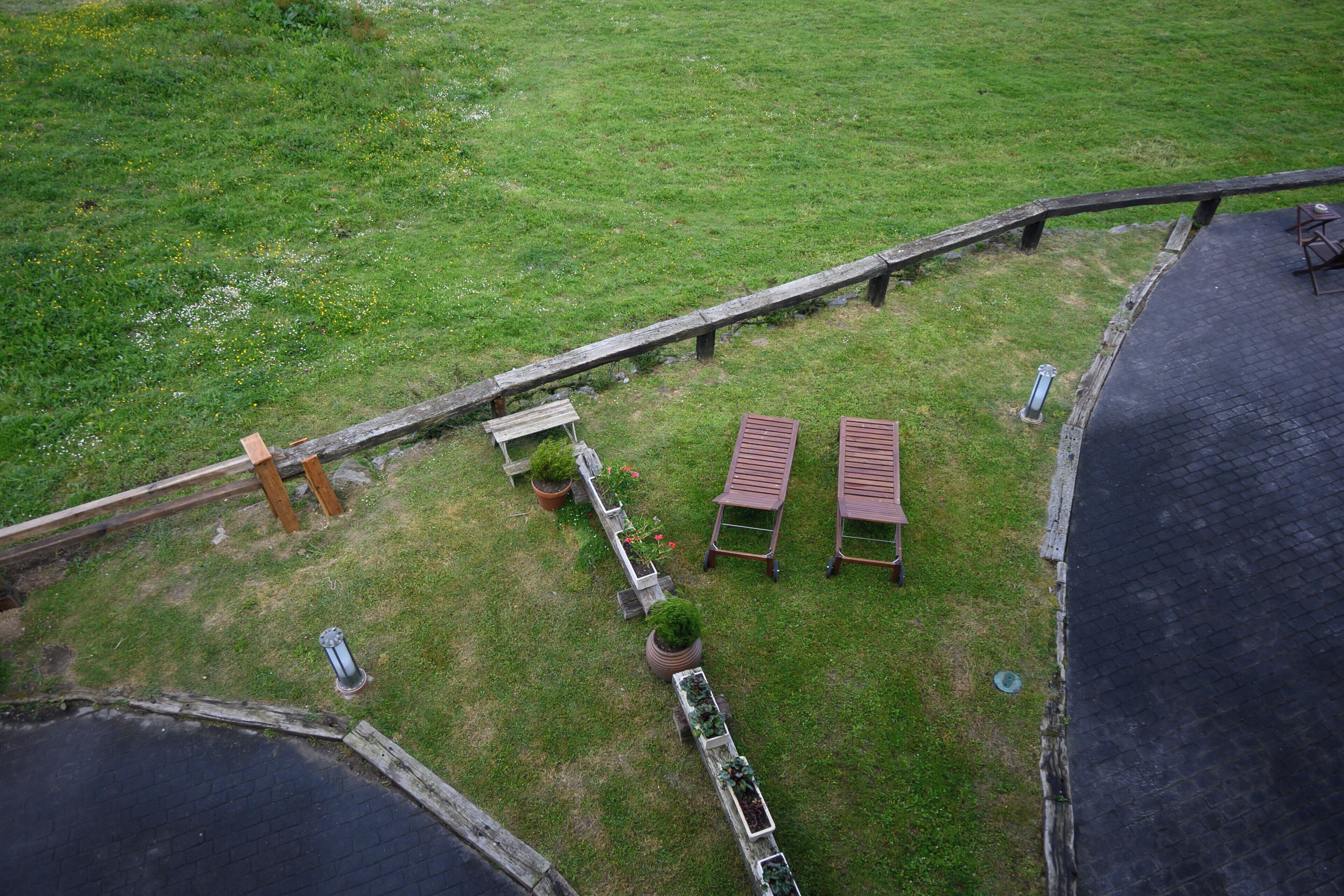 Vista aérea de una zona cubierta de césped con una terraza de madera, dos bancos y un pequeño jardín con plantas en macetas.