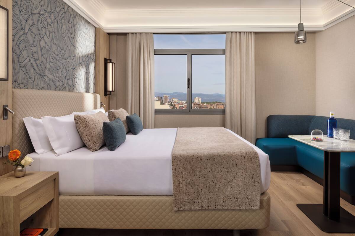 Une chambre d’hôtel comprenant un lit, un coin salon, une fenêtre donnant sur la ville et une table de chevet agrémentée d’un vase de fleurs.
