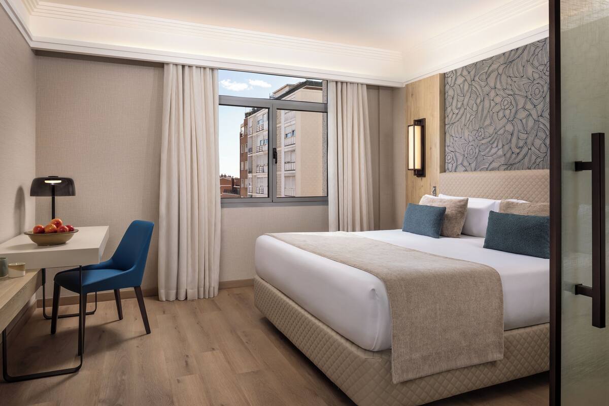 Une chambre d’hôtel comprenant un lit, un bureau avec une chaise, une fenêtre avec des rideaux et un mur orné d’un motif floral.