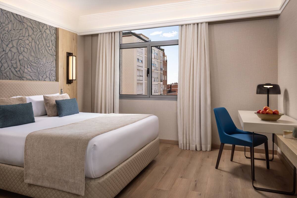 Une chambre d’hôtel avec un grand lit, un bureau avec une chaise, une fenêtre avec des rideaux et une vue sur les bâtiments environnants.