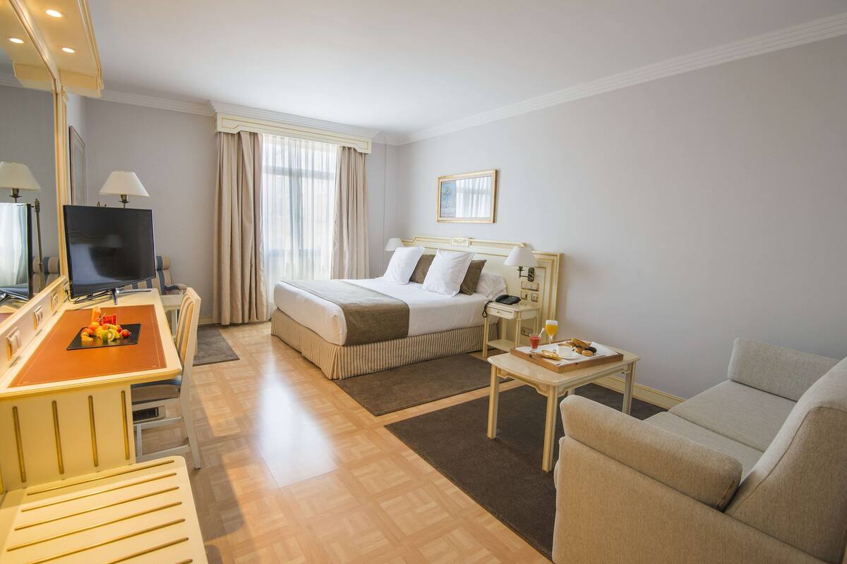 Habitación de hotel con cama, escritorio con televisor, silla, mesita con bandeja y sofá.