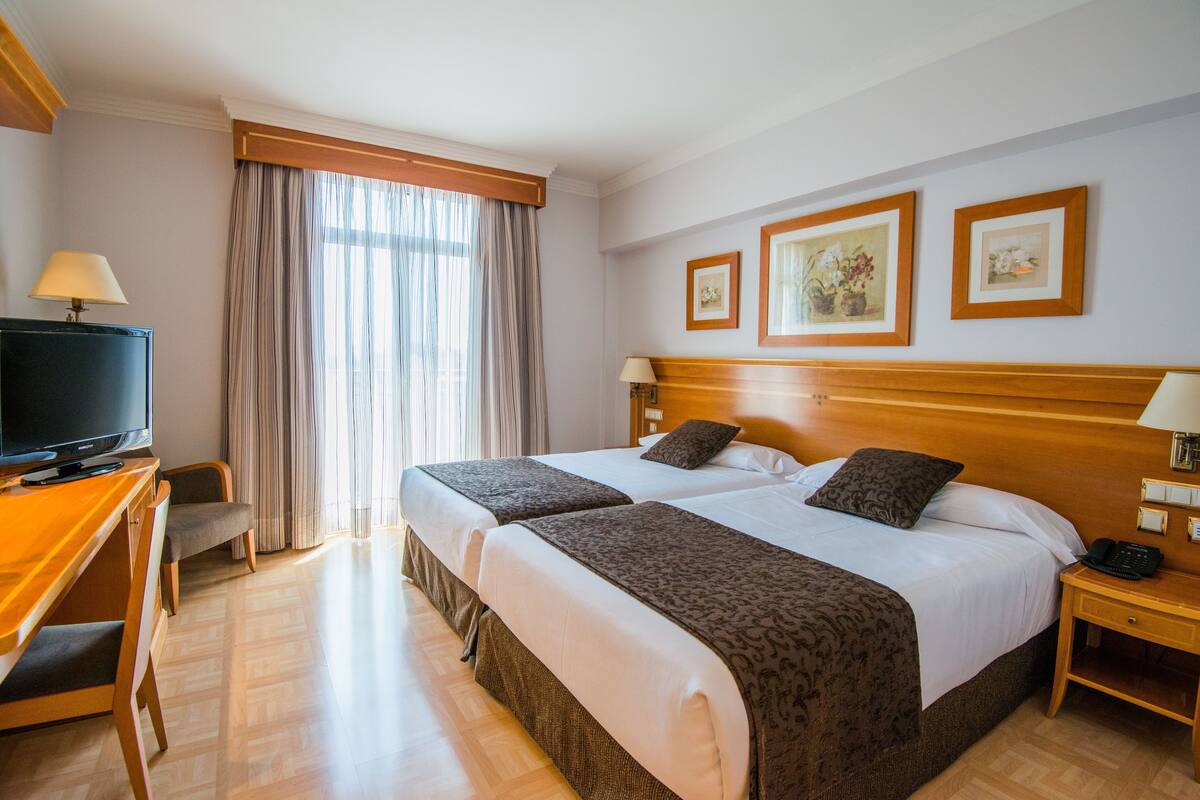 Habitación de hotel con dos camas, un escritorio con televisor, una silla, mesita de noche con lámpara y cuadros enmarcados en la pared.