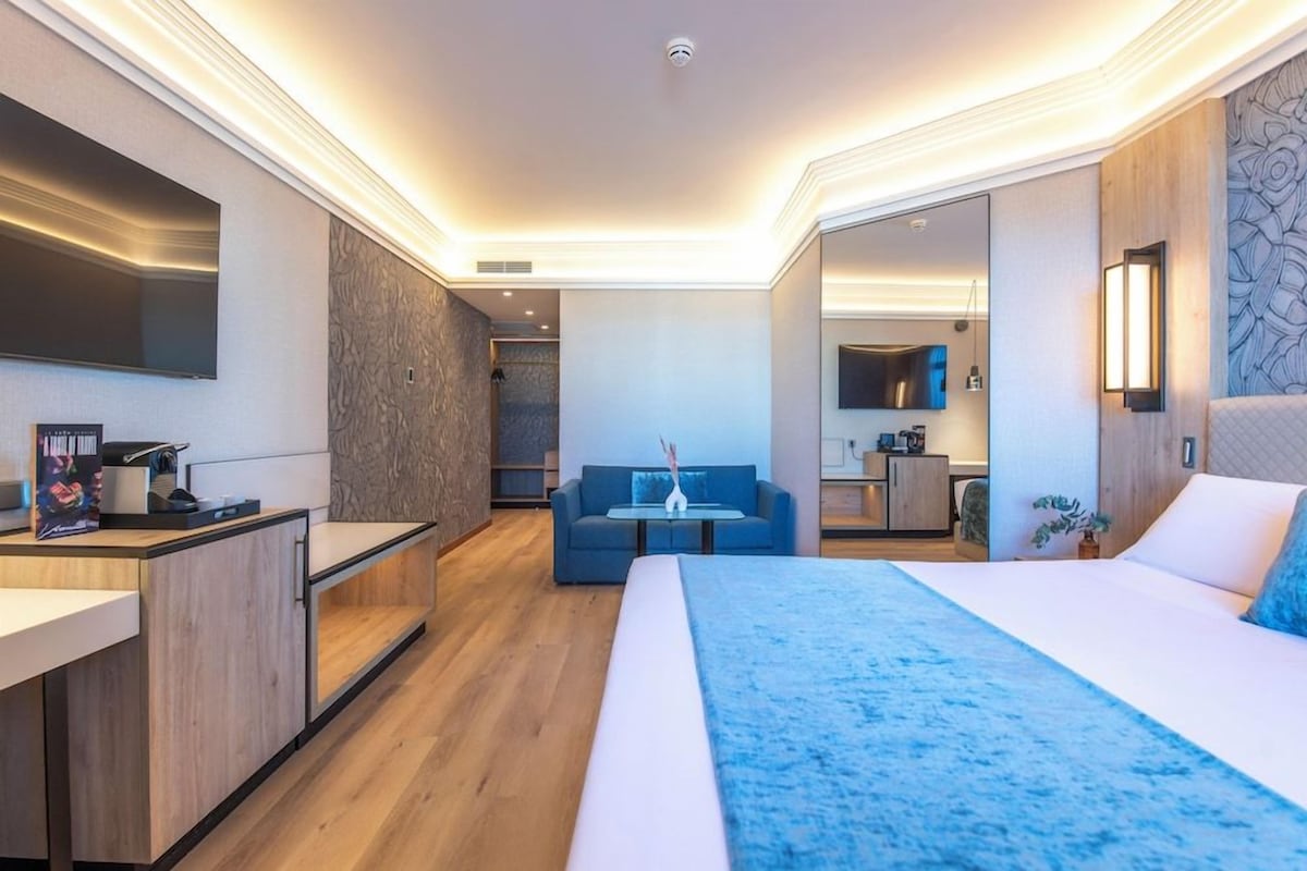 A modern hotel room with a large bed, a desk, a sofa, and a kitchenette.