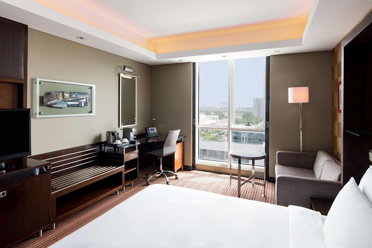 A hotel room with a bed, desk, chair, and a large window offering a city view.