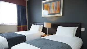 Habitación de hotel con dos camas, una mesita de noche con lámpara, un cuadro abstracto enmarcado en la pared y una ventana con cortinas.