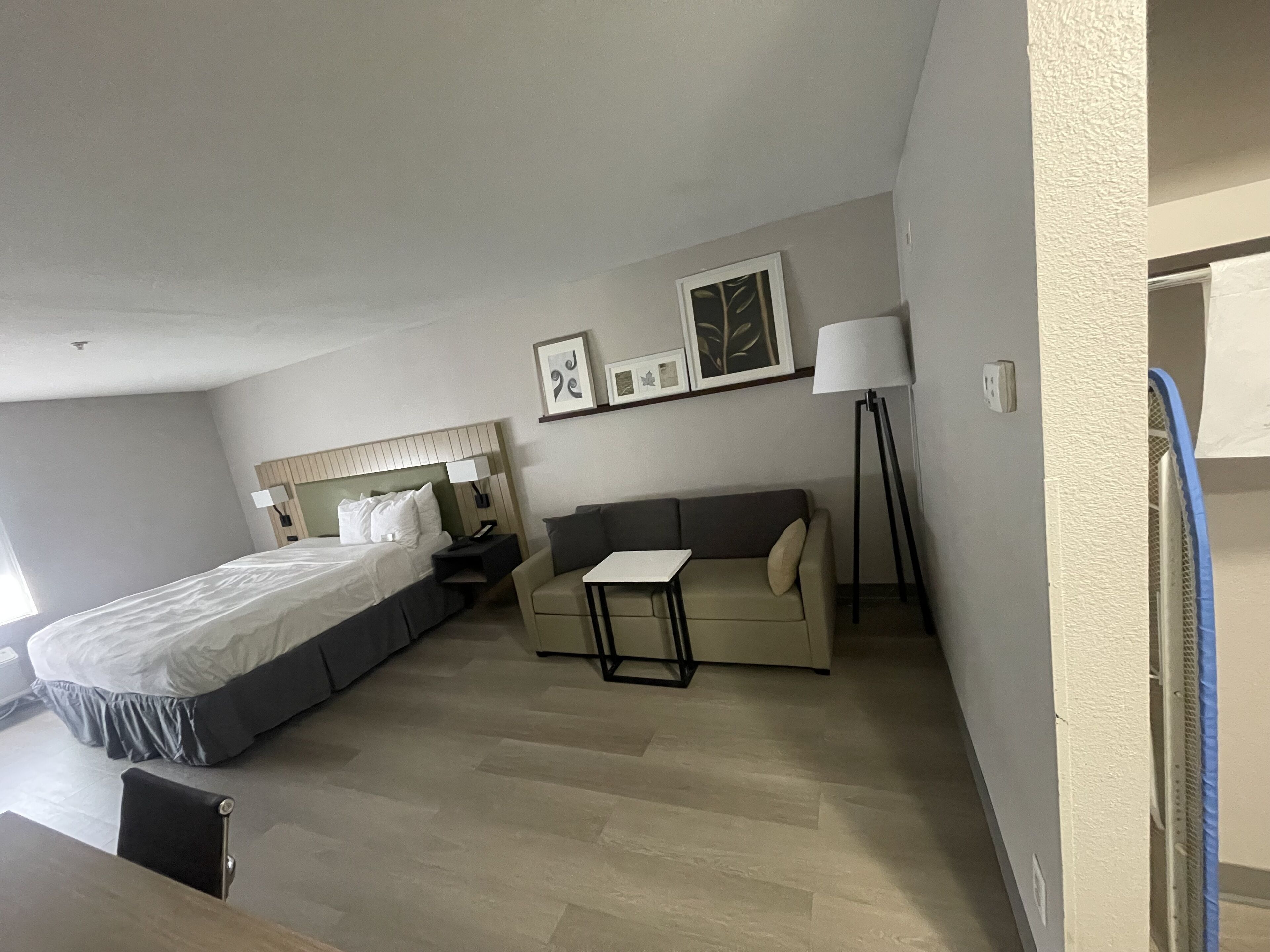 A hotel room with a bed, a sofa, a small table, a lamp, and a picture frame on the wall.