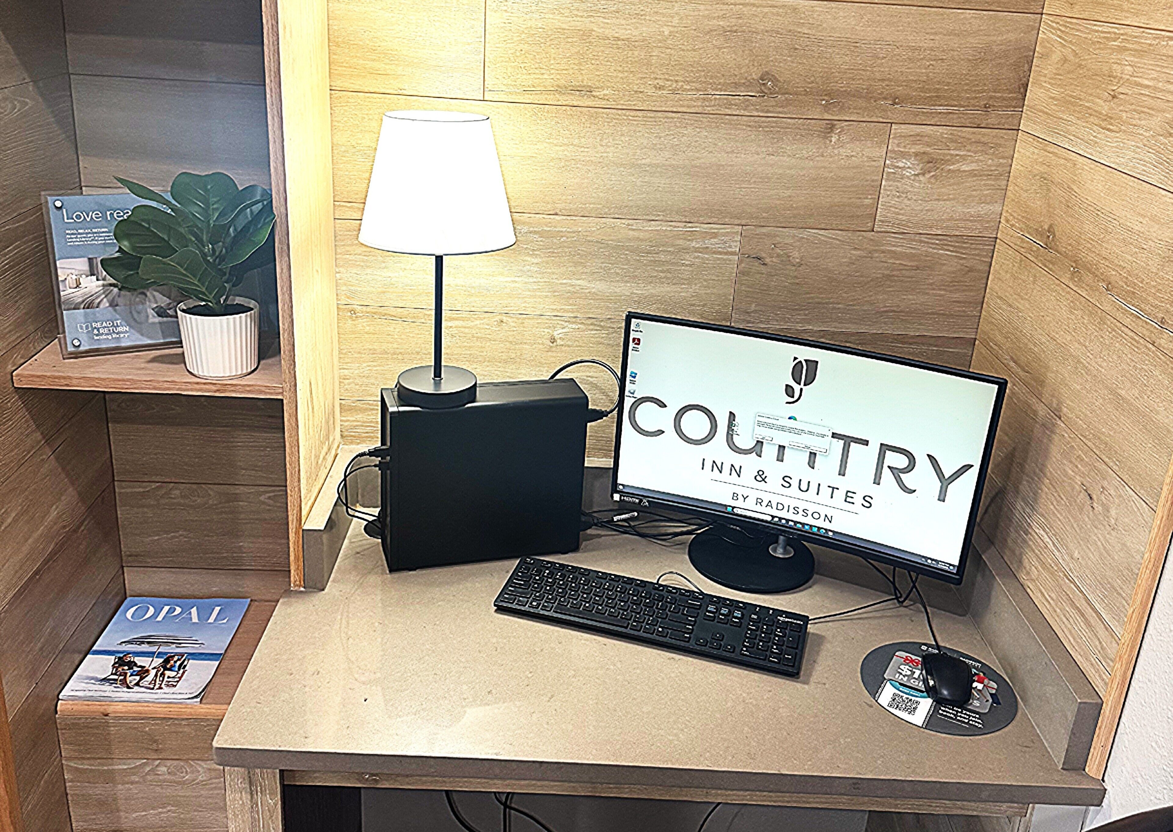 A computer desk with a monitor, keyboard, and a lamp. There is a plant on the left and a magazine titled 'OPAL' on the desk.