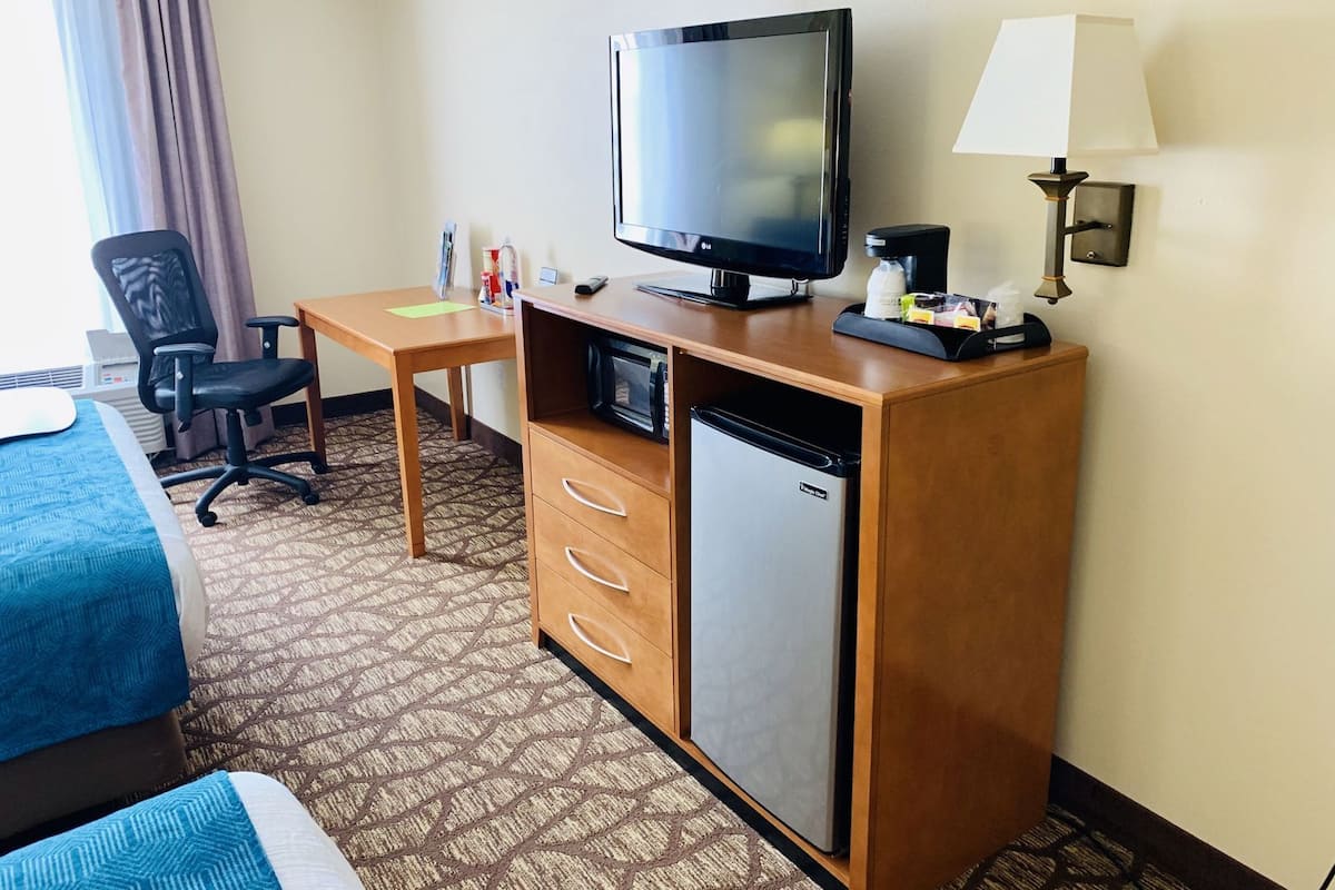Una habitación de hotel con cama, escritorio con televisor, refrigerador, silla y lámpara.