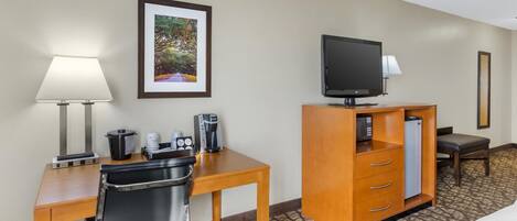 A hotel room with a wooden desk, a chair, a television on a stand, a coffee maker, a refrigerator, and a bed.
