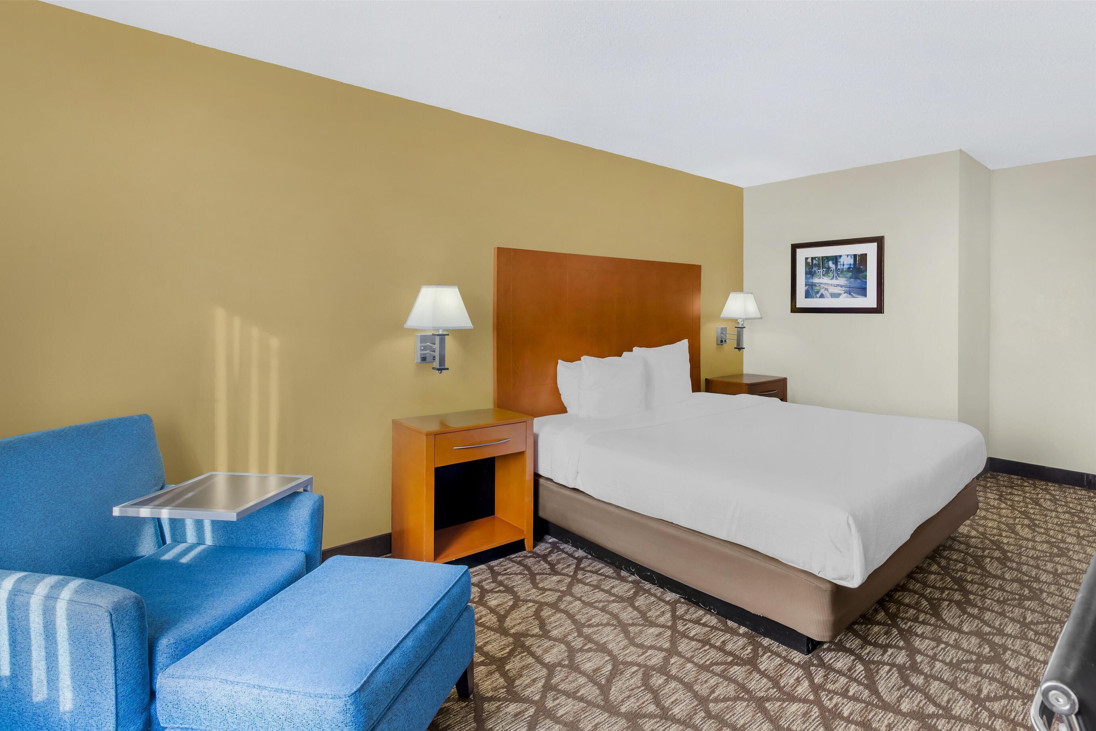 A hotel room with a bed, a blue armchair, a small table, and a nightstand with lamps.