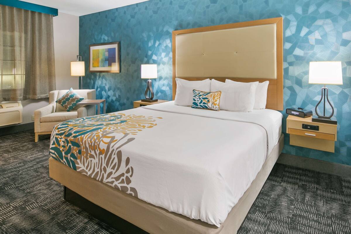 A hotel room with a large bed, two bedside tables, a chair, a desk, and a wall with a geometric pattern.