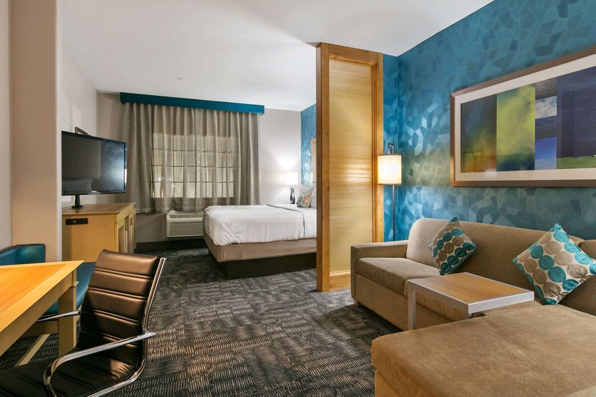 A hotel room with a bed, a sofa, a desk with a chair, a TV, and a painting on the wall.