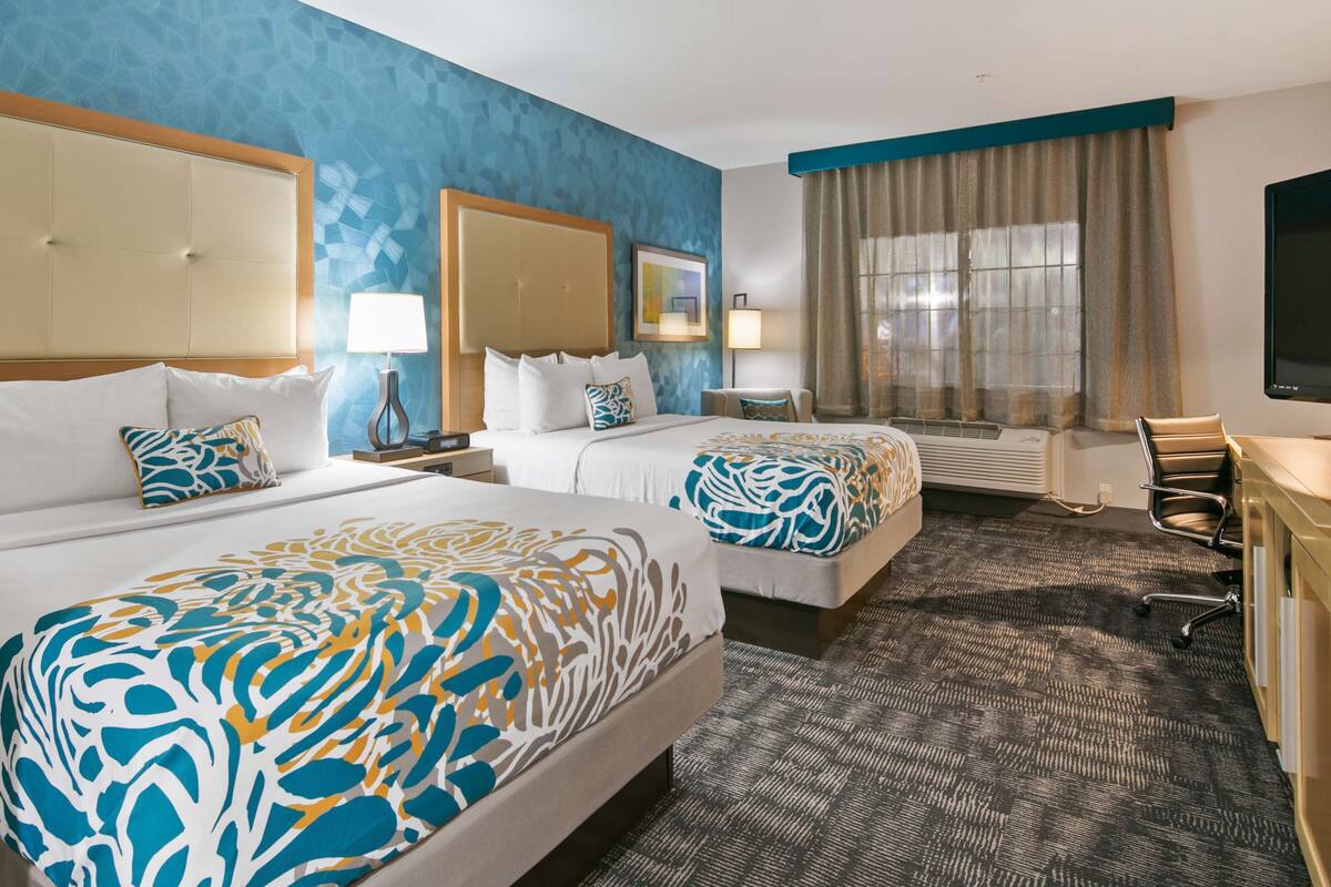 A hotel room with two beds, a desk, a TV, and a large window with curtains.
