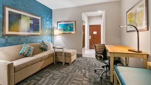 A hotel room with a beige sofa, a small table, a desk with a chair, and a patterned carpet. - Best Western Plus Houston Atascocita Inn & Suites (Humble)