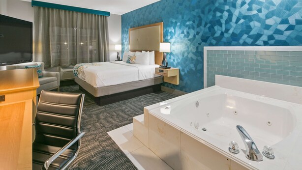 A hotel room with a large bed, a sitting area with a chair, a desk, a TV, a bathroom with a bathtub, and a wall with a geometric pattern. - Best Western Plus Houston Atascocita Inn & Suites (Humble)