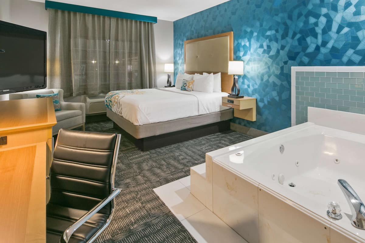 A hotel room with a large bed, a sitting area with a chair, a desk, a TV, a bathroom with a bathtub, and a wall with a geometric pattern.