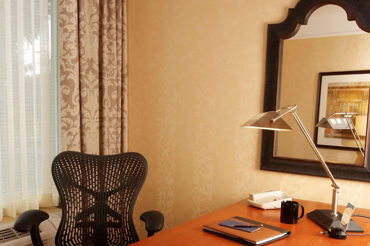 A hotel room with a wooden desk, a black office chair, a desk lamp, a framed picture on the wall, and a window with curtains.