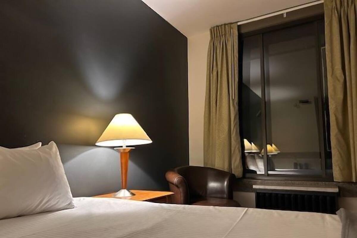 A hotel room with a bed, a nightstand with a lamp, a leather armchair, and a window with curtains.