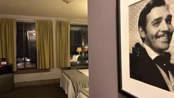 Quarto de hotel com cama, escrivaninha, cadeira, luminária e janela com cortinas.