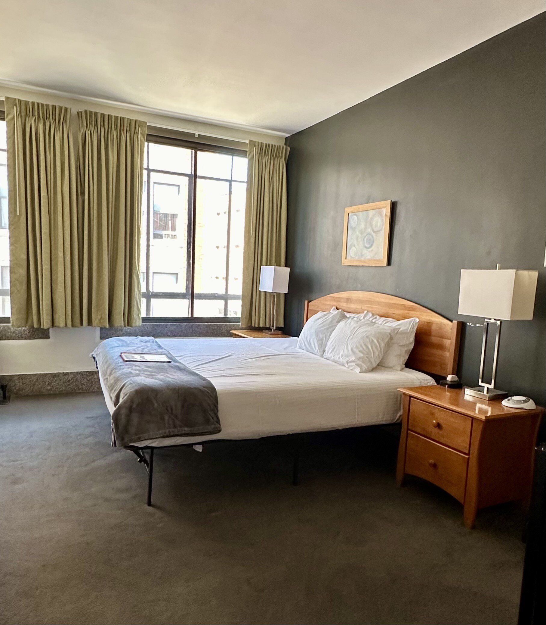 Una habitación de hotel con cama, mesitas de noche, una ventana con cortinas y una lámpara de pared.
