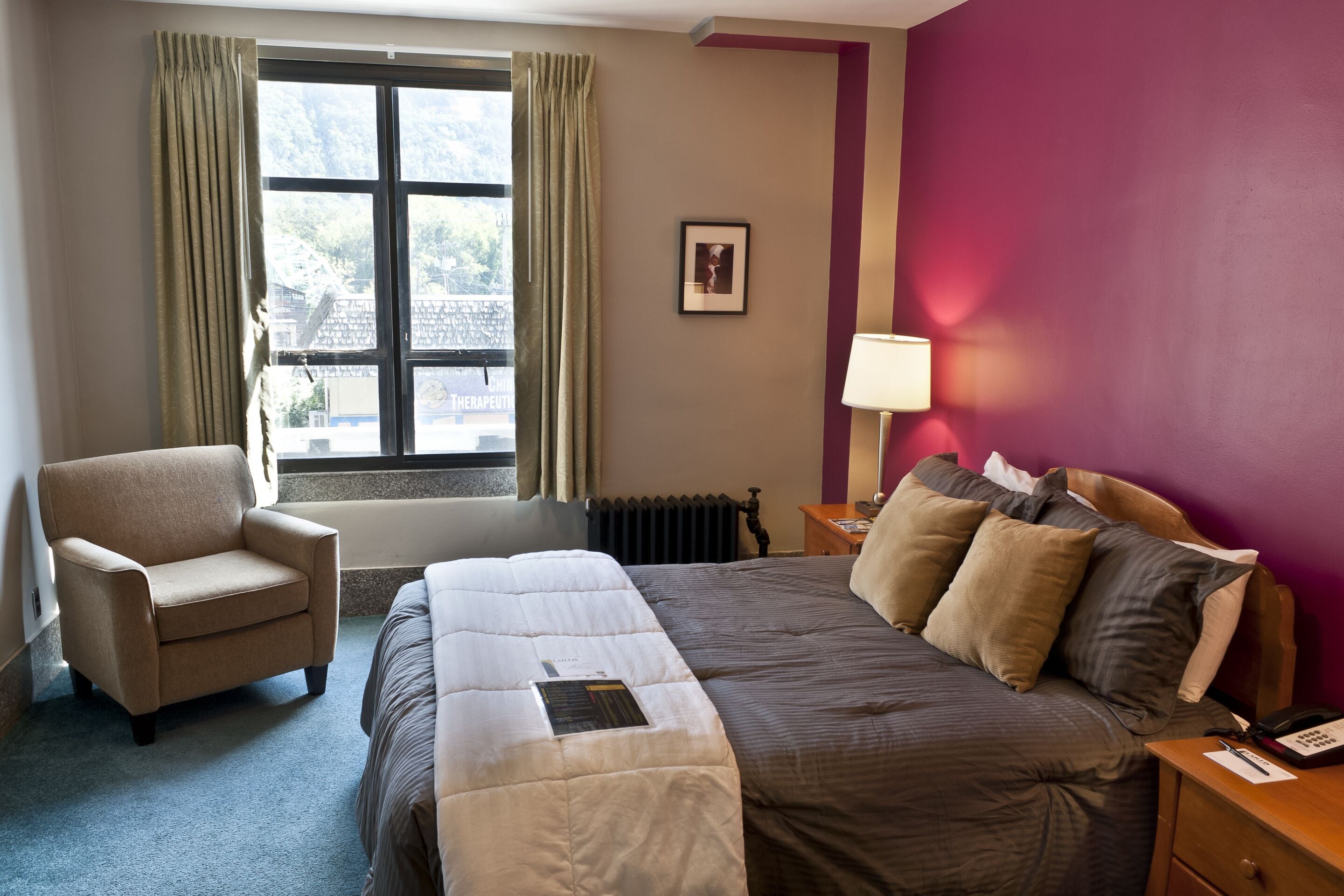 Een hotelkamer met een bed, een stoel, een raam met gordijnen, een radiator, een nachtkastje met een lamp, en een lijst met een afbeelding aan de muur.