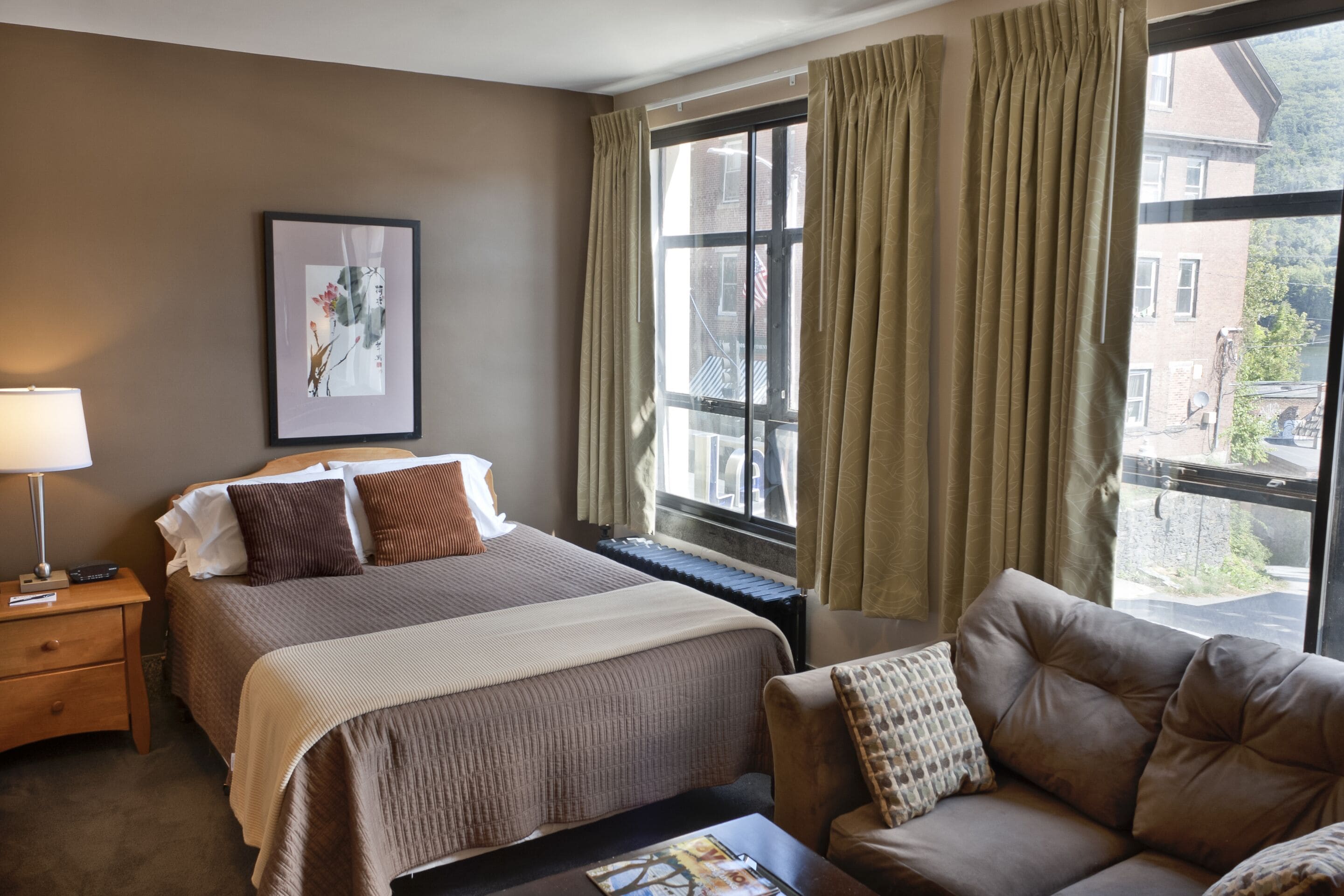 A hotel room with a bed, a sofa, a window with curtains, a framed picture on the wall, and a nightstand with a lamp.