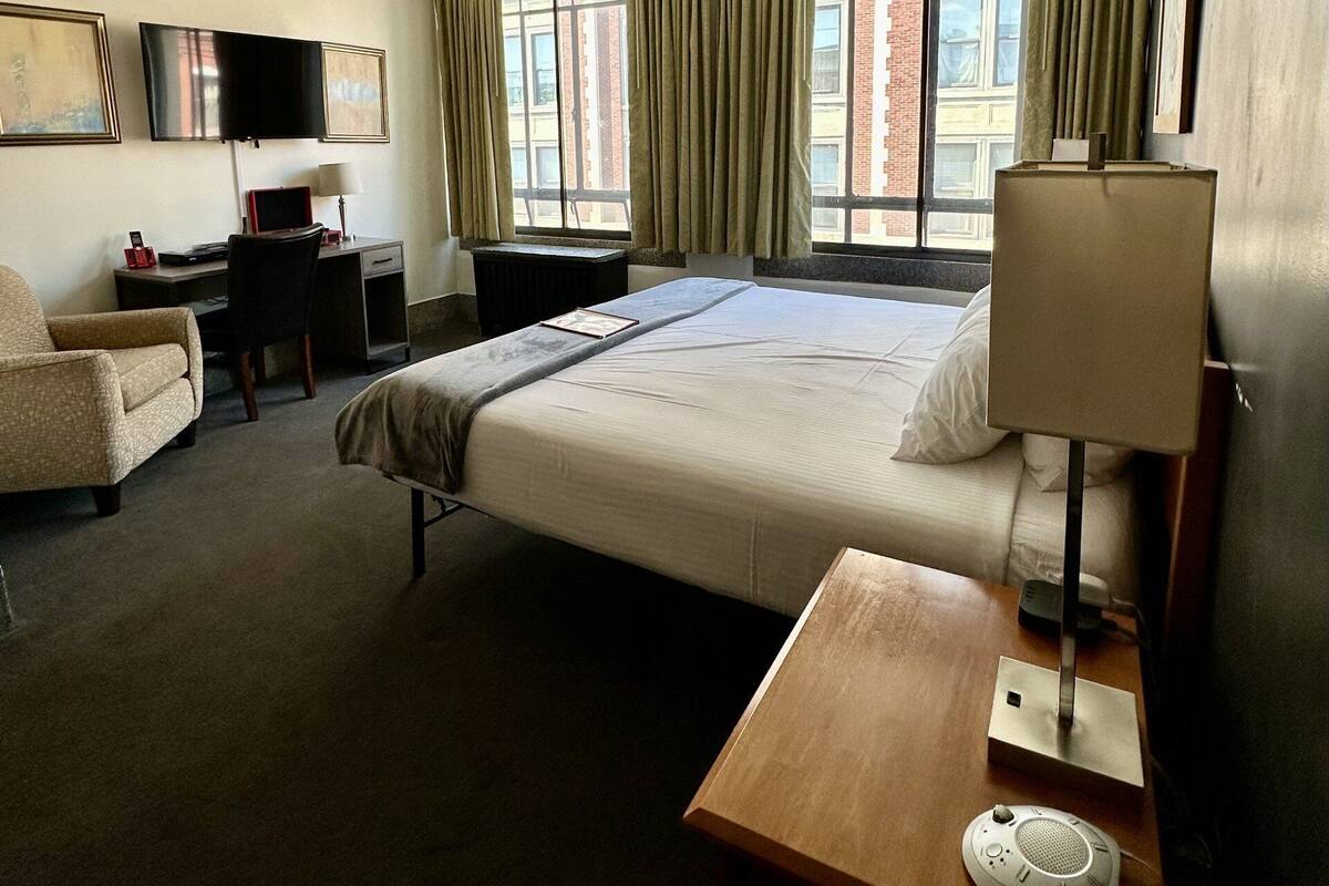 A hotel room with a bed, a desk, a chair, a TV, and a window with curtains.