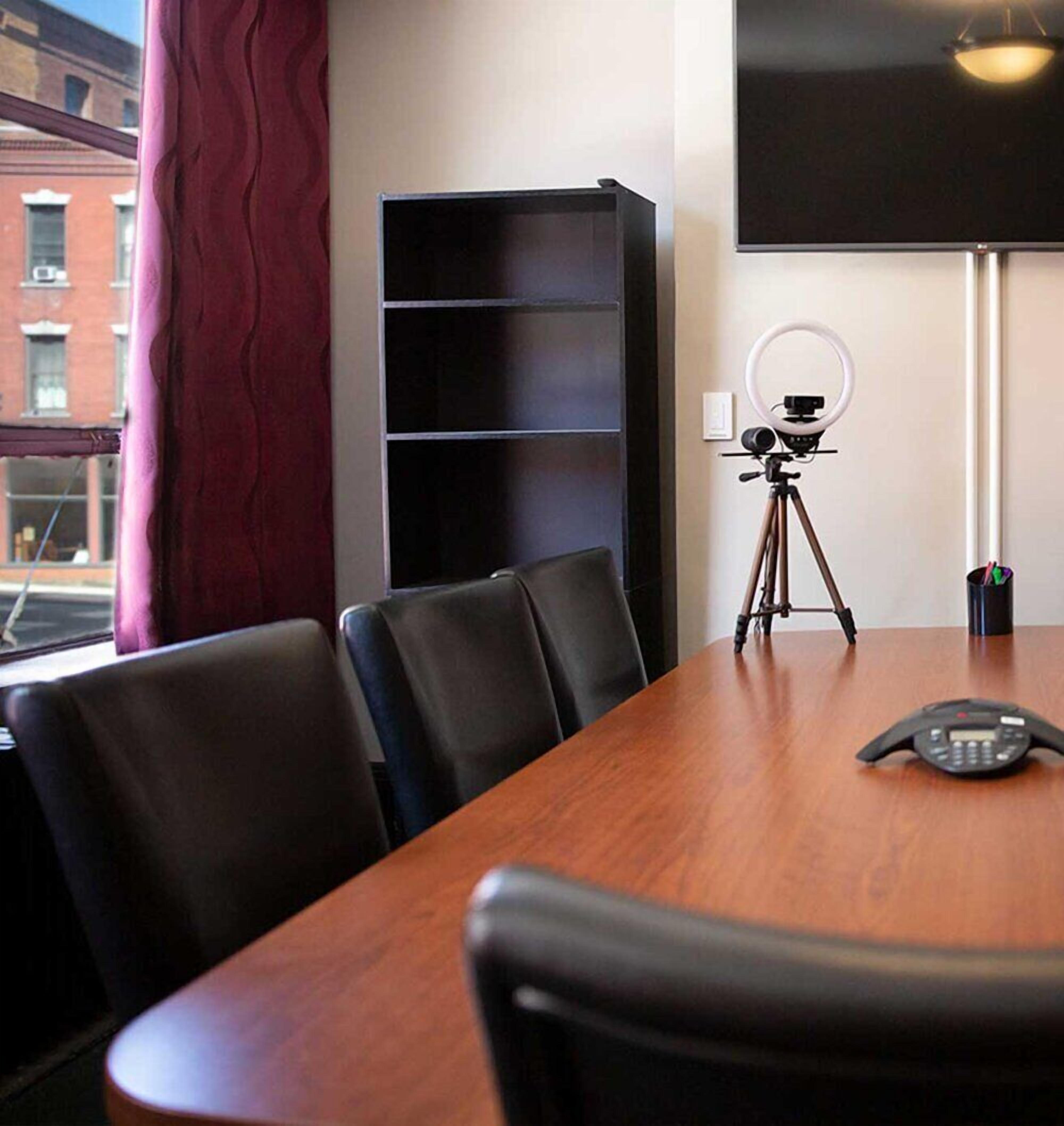 Un salón de conferencias con una mesa de madera, sillas negras, una pantalla de proyector, una estantería y una luz de aro sobre un trípode.