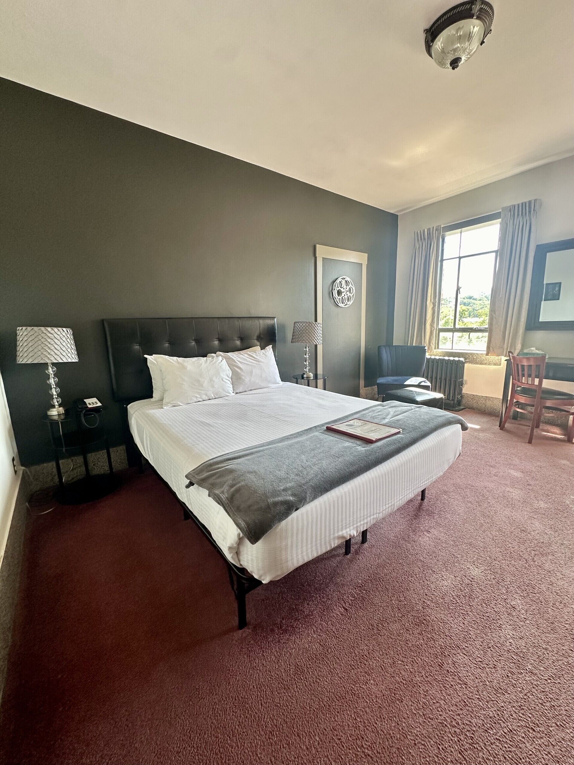 A hotel room with a large bed, a bedside table with a lamp, a chair, and a window with curtains.
