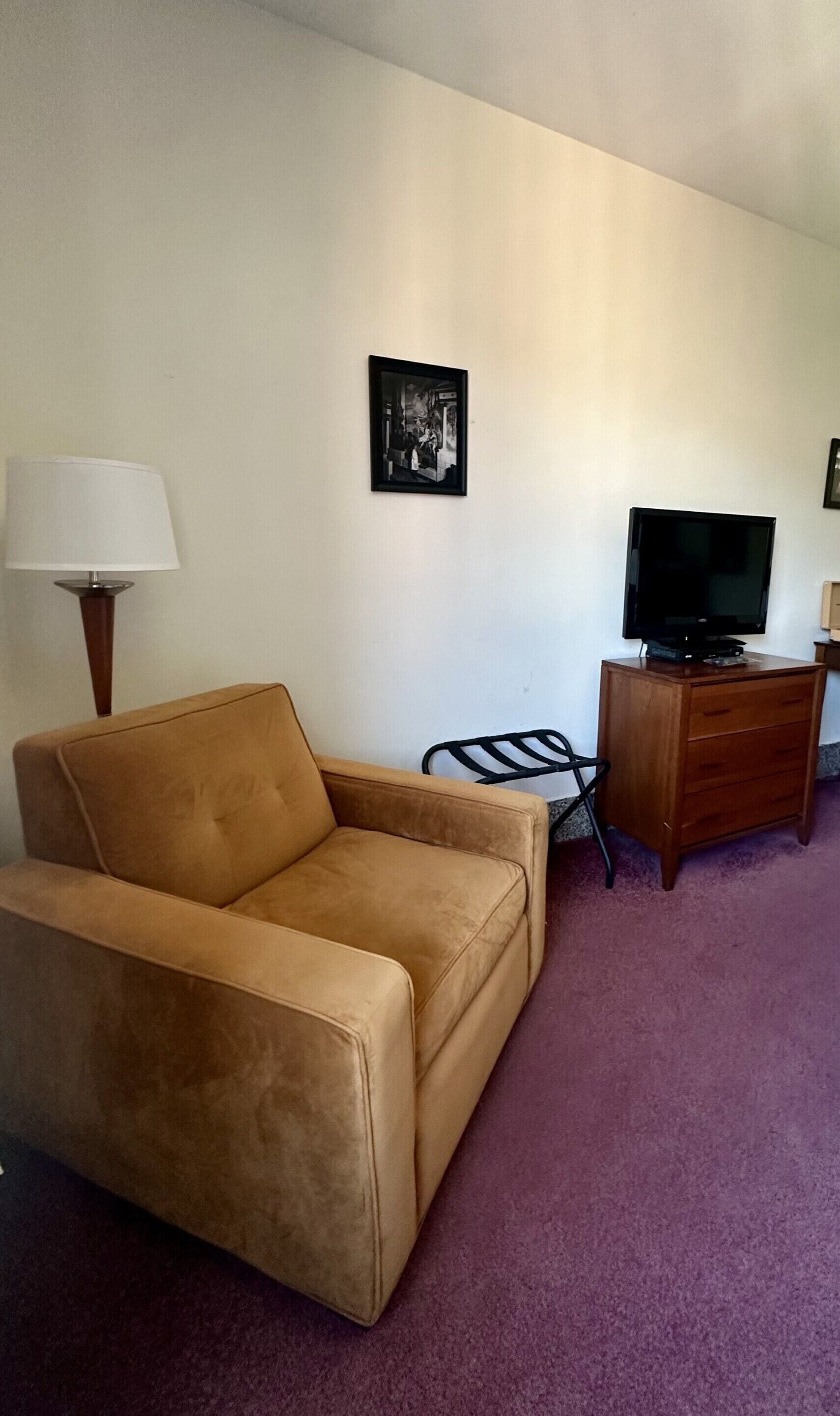 Ein Wohnzimmer mit einem beigefarbenen Sofa, einem Fernseher auf einer Holzkommode und einer Lampe.