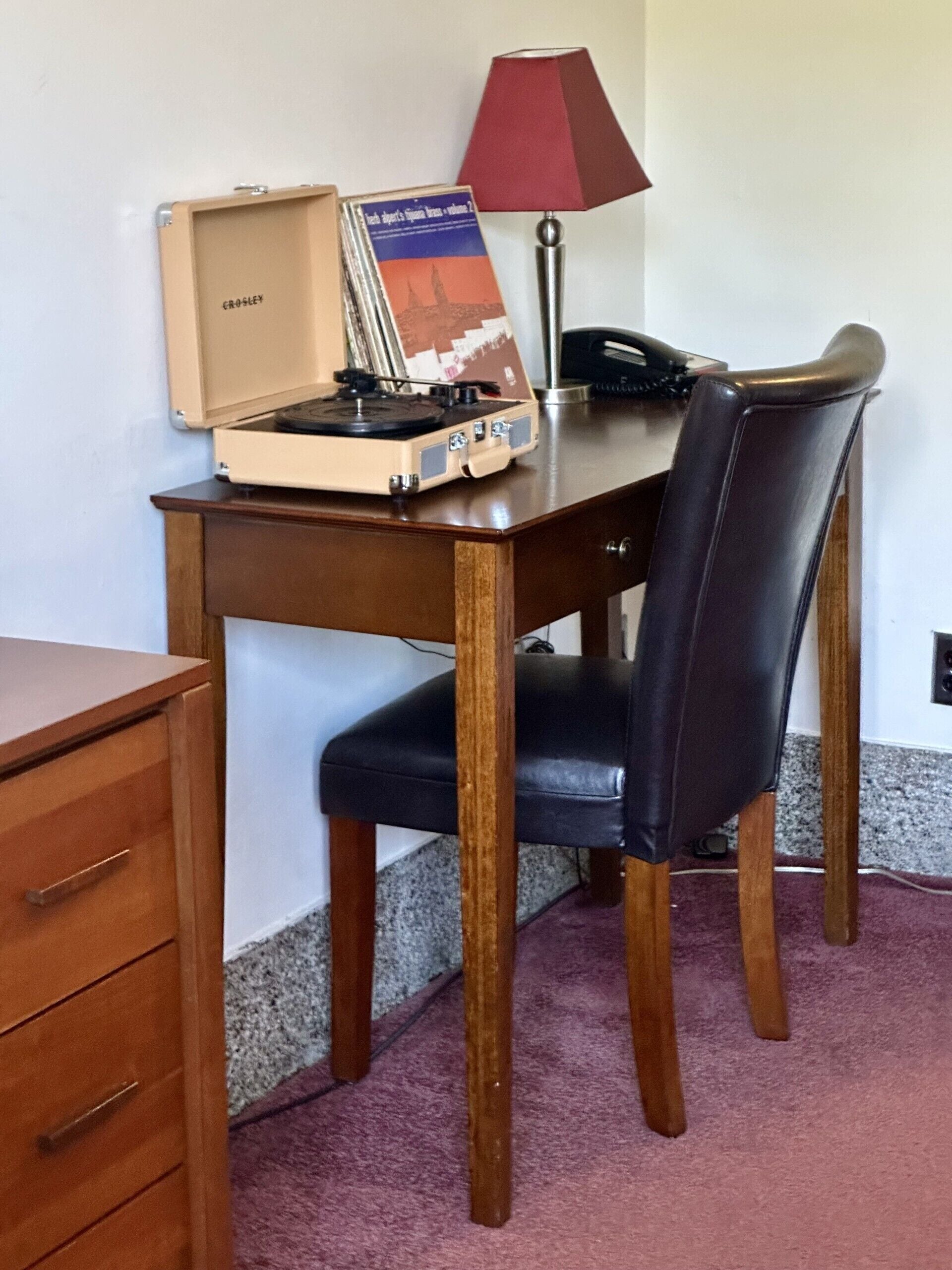 A wooden desk with a vintage record player, a red lamp, and a black leather chair.