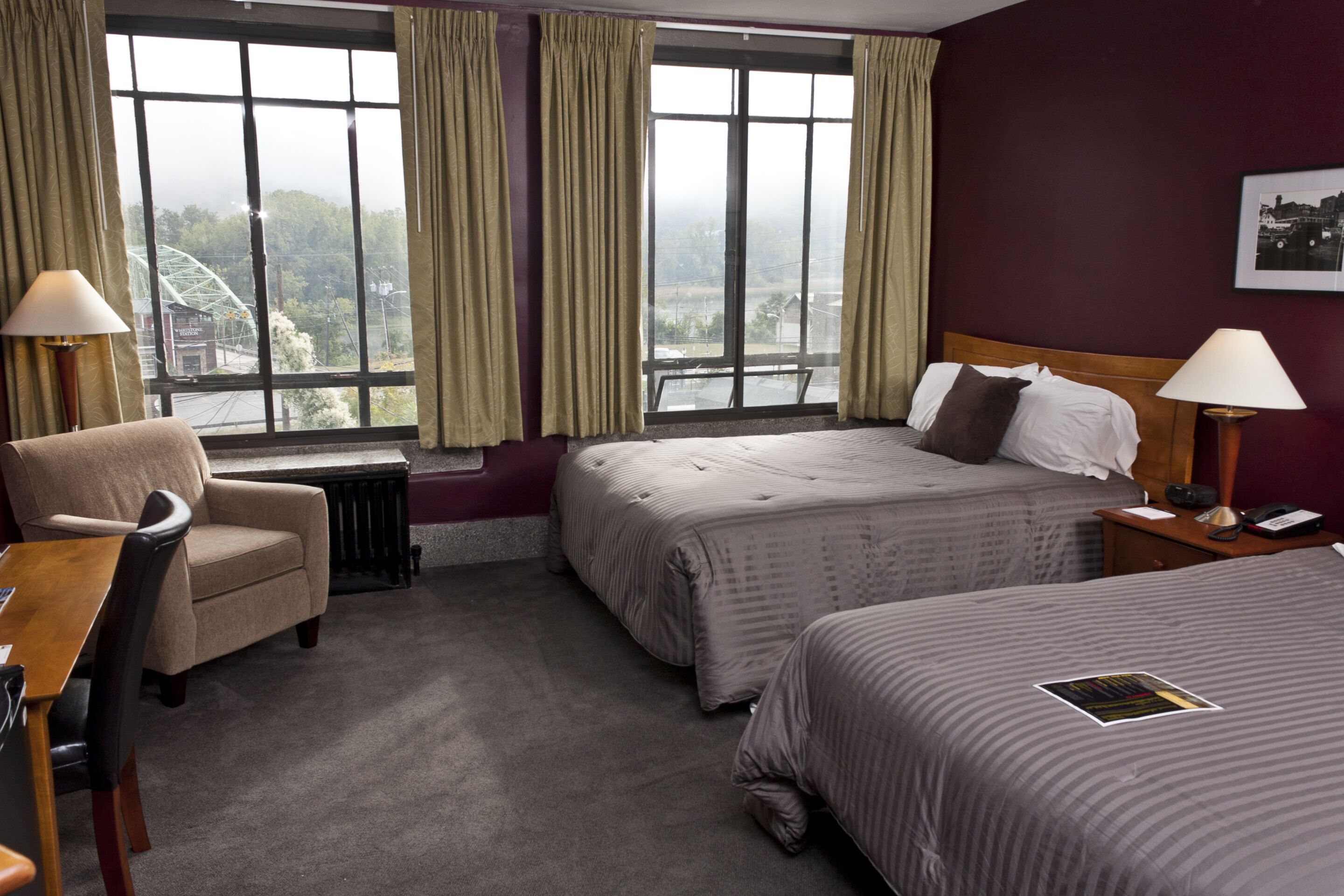 A hotel room with two beds, a desk, and a chair. There are large windows with curtains, a radiator, and a lamp on the desk.