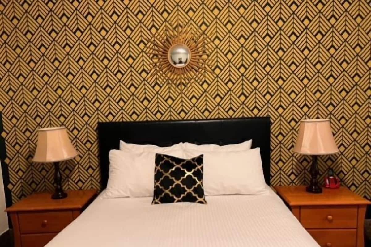 A neatly made bed with white linens and a black pillow with a gold pattern. There are two bedside tables with lamps on either side of the bed.