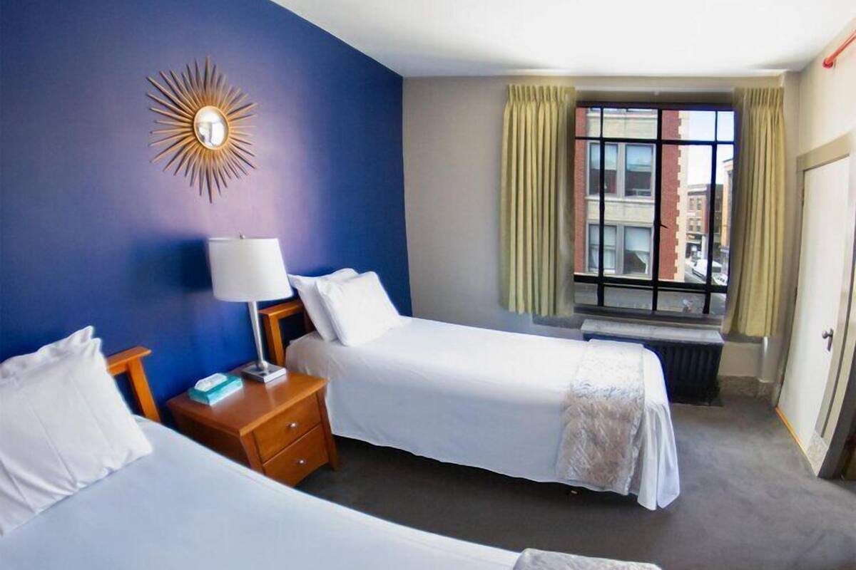 A hotel room with two single beds, a bedside table with a lamp, a window with curtains, and a wall-mounted sunburst light fixture.
