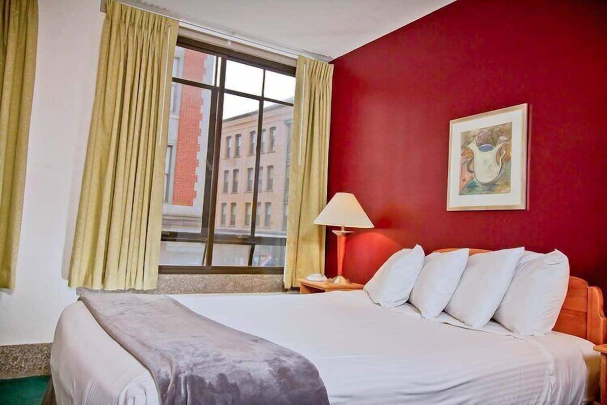A hotel room with a bed, a nightstand, a lamp, a window with curtains, and a painting on the wall.