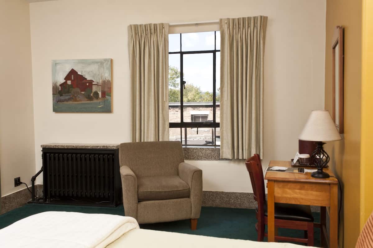 A hotel room with a bed, a chair, a table with a lamp, a radiator, a window with curtains, and a painting on the wall.