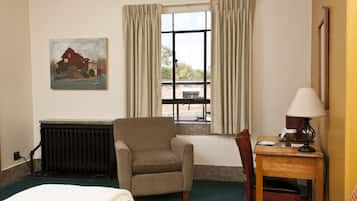 Een hotelkamer met een bed, een stoel, een tafel met een lamp, een radiator, een raam met gordijnen en een schilderij aan de muur.