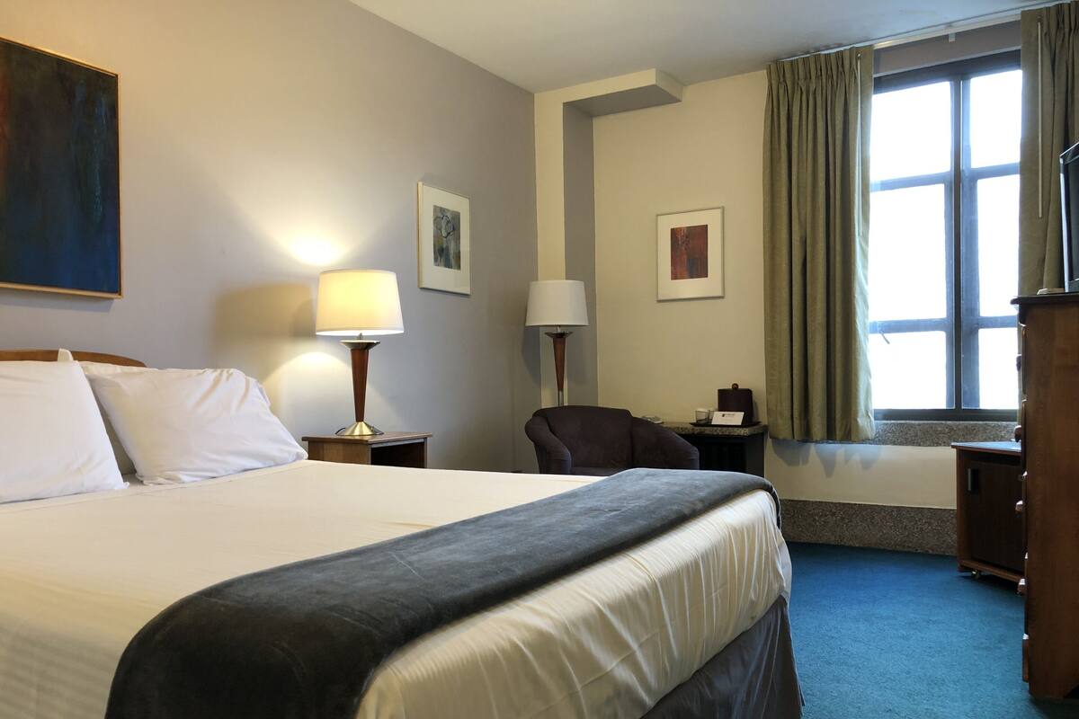 A hotel room with a large bed, two bedside tables with lamps, a chair, a desk with a lamp, and a window with curtains.