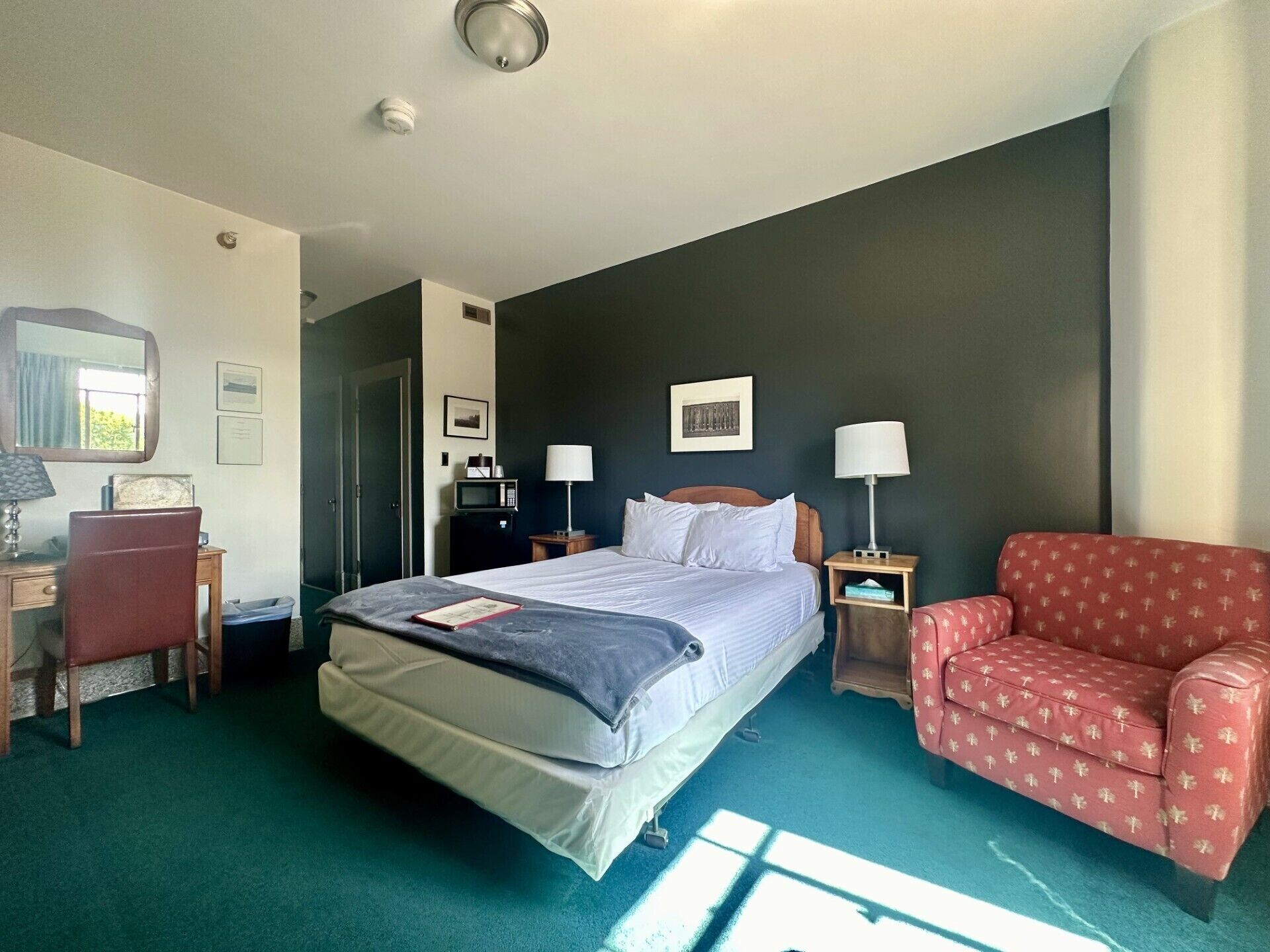 A hotel room with a bed, a desk, a chair, a red patterned sofa, and a microwave on a bedside table.