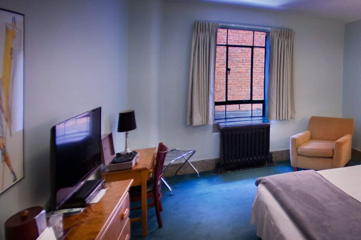A hotel room with a bed, a desk with a computer, a chair, a window with curtains, a radiator, and a lamp.