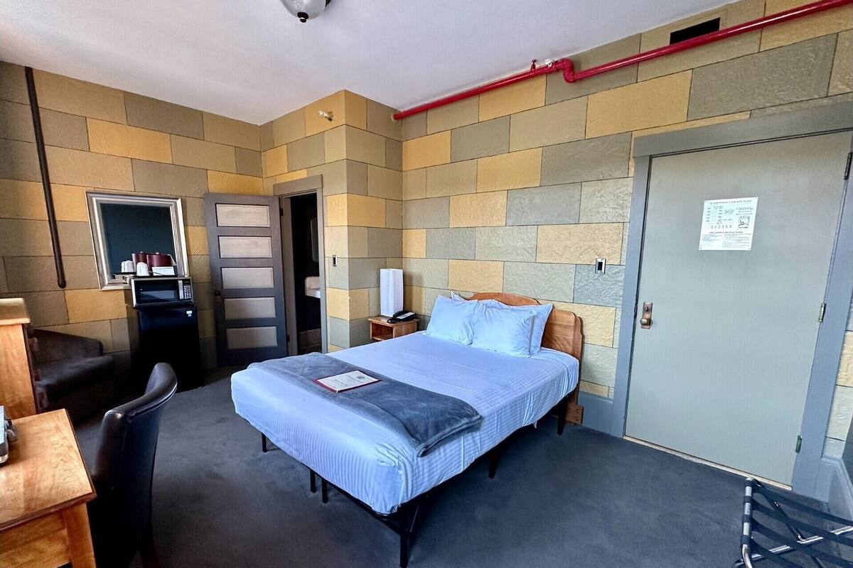 A hotel room with a bed, a desk with a chair, a microwave, a telephone, and a door with a sign.
