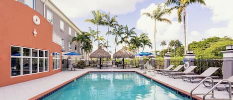A swimming pool with lounge chairs and umbrellas, surrounded by palm trees and a building with a red facade.