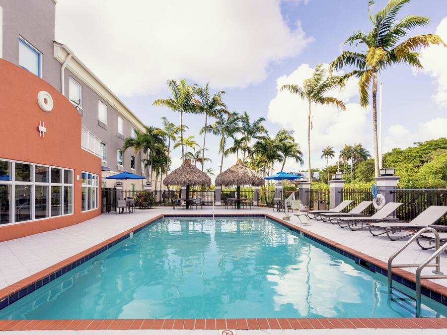 A swimming pool with lounge chairs and umbrellas, surrounded by palm trees and a building with a red facade.