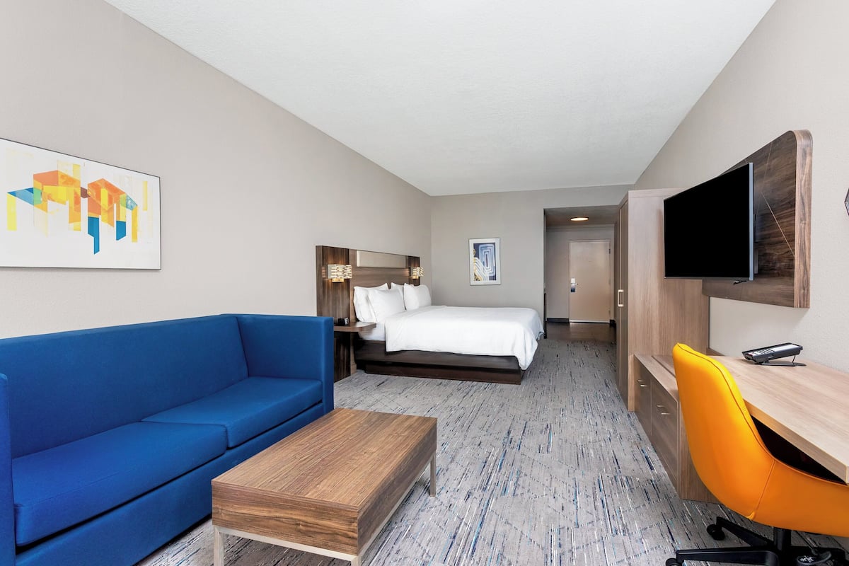 A hotel room with a blue sofa, a wooden coffee table, a bed with white bedding, a desk with a chair, and a flat-screen TV mounted on the wall.